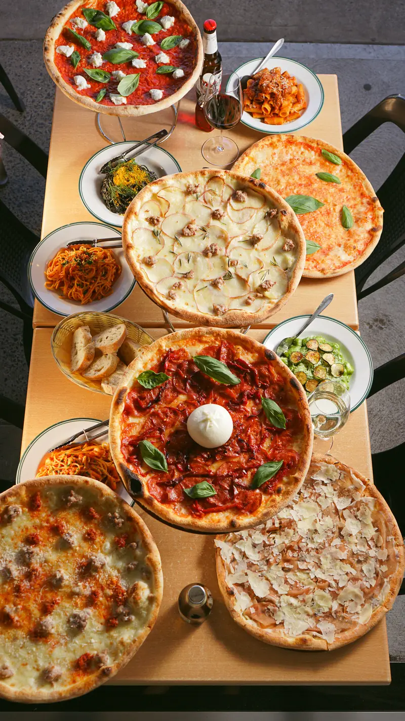 A long table from above: six pizzas, pasta, salads and bread